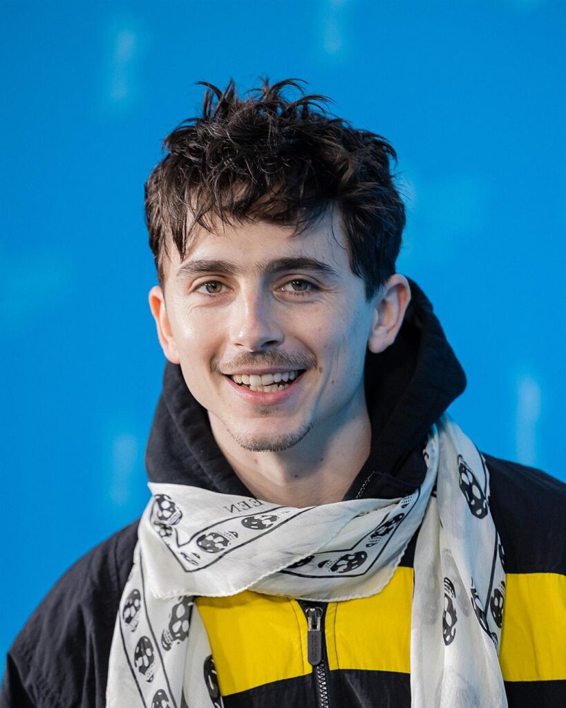 Timothée Chalamet smiling at a photo call, in a black and yellow jacket and a white scarf