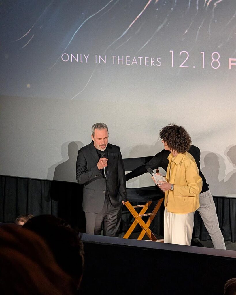 Denis Villeneuve speaking on stage at a Dune trailer launch, with the 12.18 release date on the screen