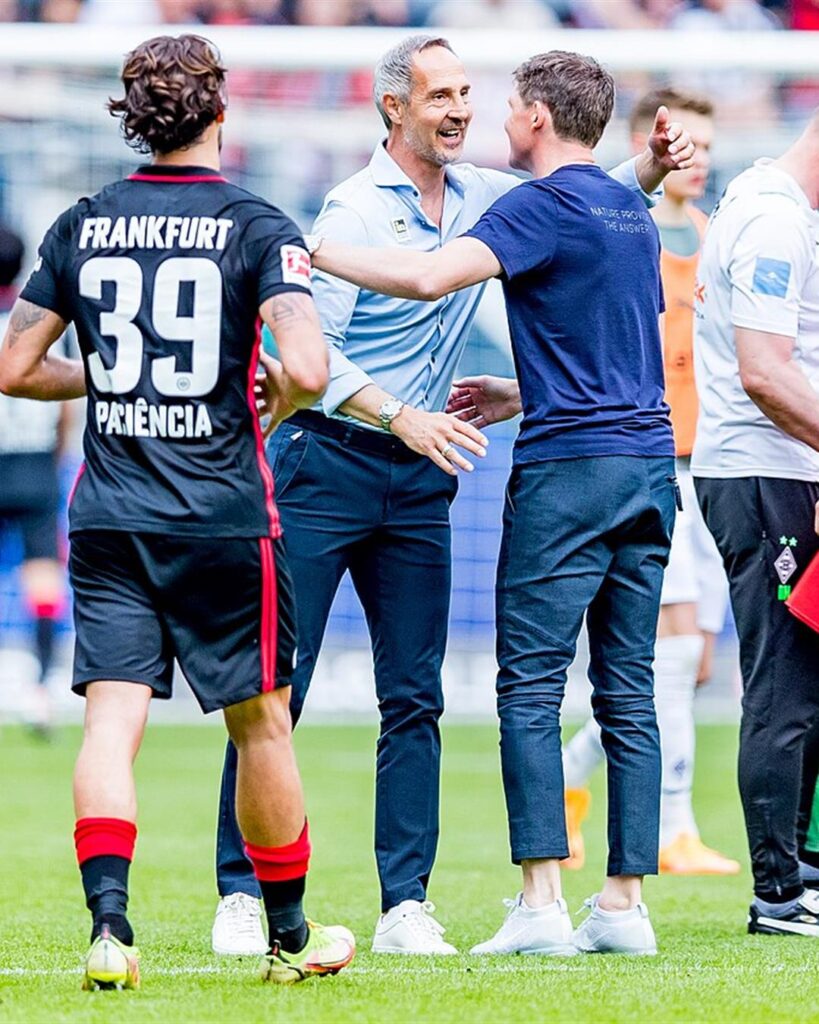Adi Hütter smiling and hugging a coach on the pitch after a Bundesliga game in Frankfurt