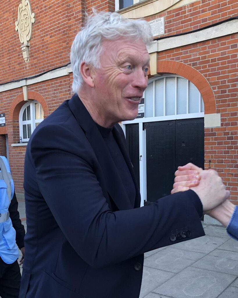 David Moyes in a dark jacket, smiling and shaking hands with a fan outside Craven Cottage