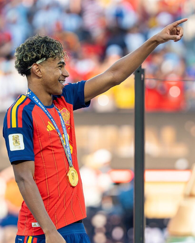 Lamine Yamal smiling and pointing to the crowd, wearing a gold medal and a red Spain shirt
