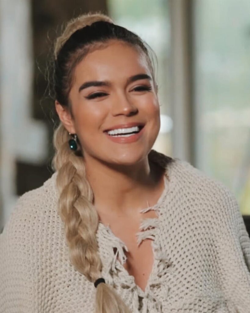 Karol G laughing during an interview, with a long blonde braid and a cream sweater
