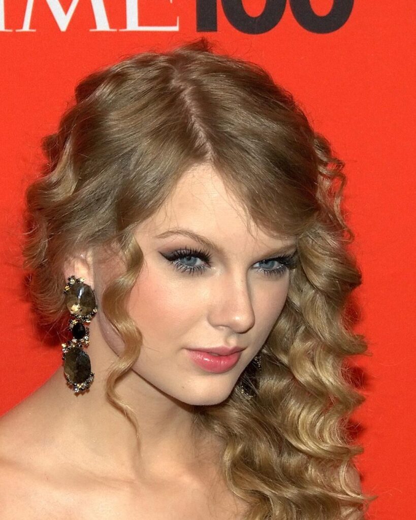 Taylor Swift with long curly hair and large earrings in front of a red backdrop at the 2010 Time 100 event