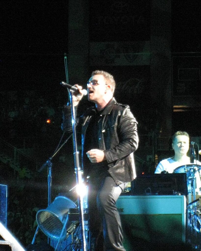 Bono in sunglasses and a black leather jacket singing on stage, with drummer Larry Mullen Jr behind him, 2009